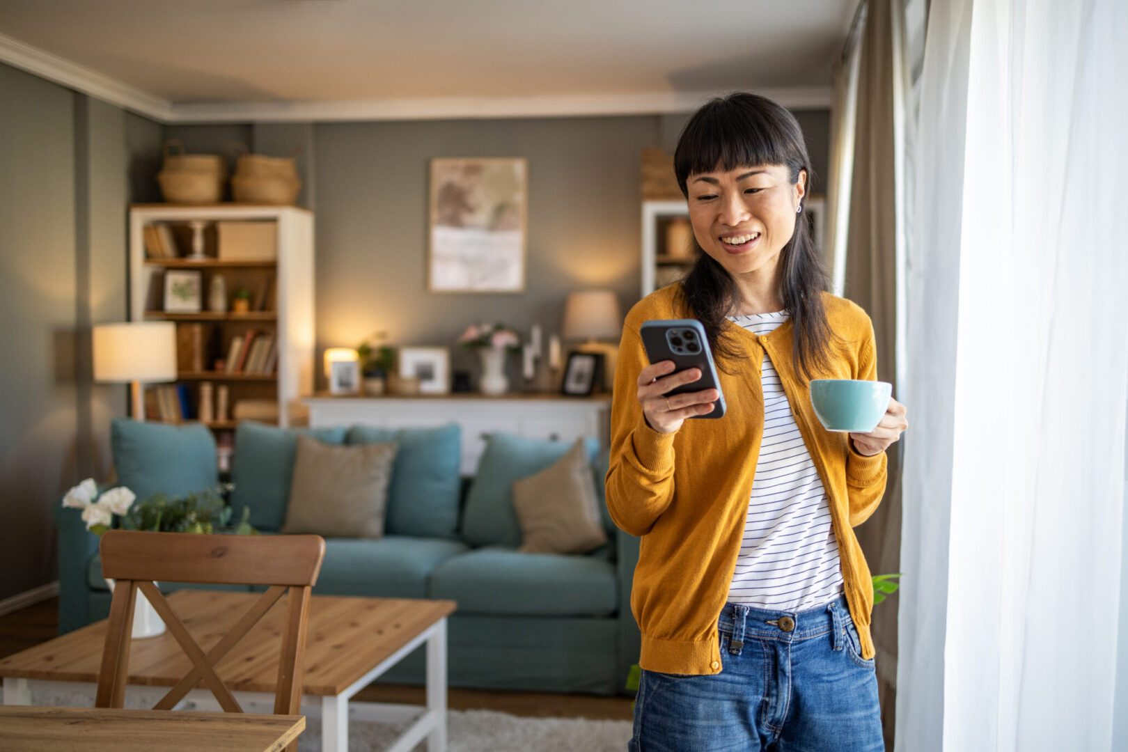 Personne debout dans un salon lumineux tenant un smartphone et une tasse près d’une fenêtre.