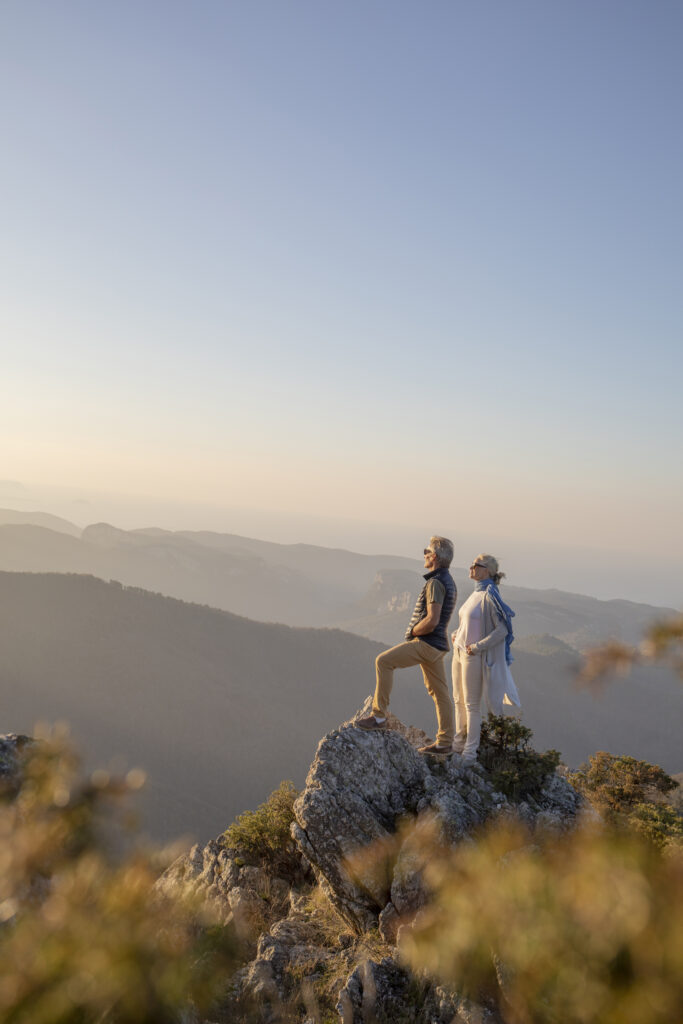 Deux personnes debout sur un sommet rocheux face à un paysage de montagnes au coucher du soleil.