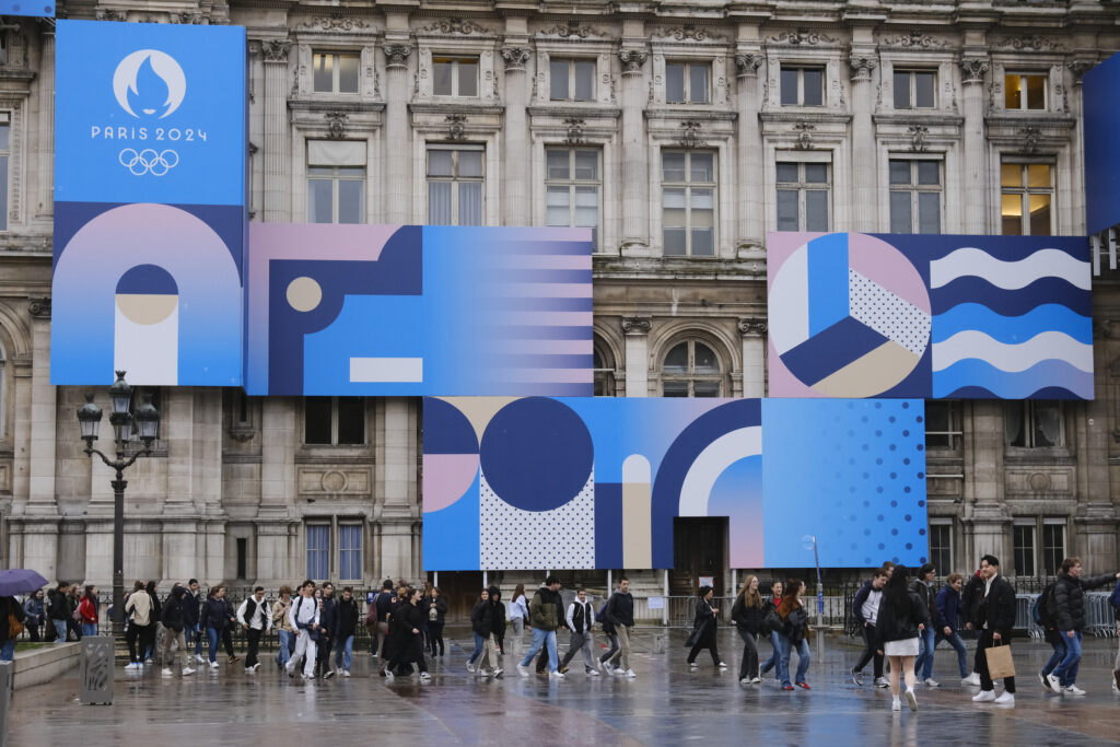 Façade d’un bâtiment institutionnel habillée d’éléments graphiques Paris 2024, avec circulation de passants sur une place urbaine.